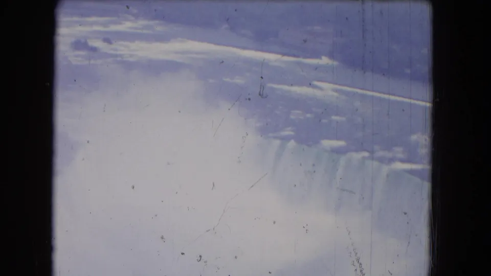 1964 Niagara Falls frame showing the cascade edge and misty lower area, with soft exposure, grain, gate edges, and scratches from the 8mm source.