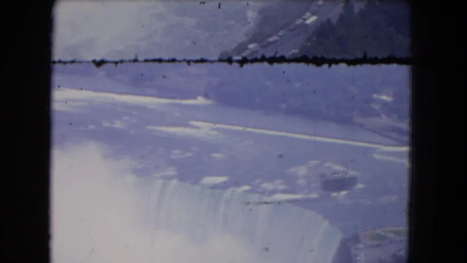1964 Niagara Falls aerial frame with the river, snowy or icy banks, the waterfall rim, mist, and visible 8mm frame damage.