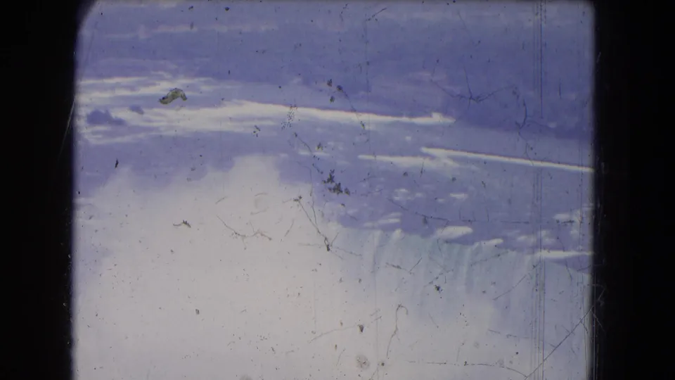 1964 8mm frame showing Niagara Falls, heavy mist, distant shoreline, and surface scratches across a faded travel-film view.