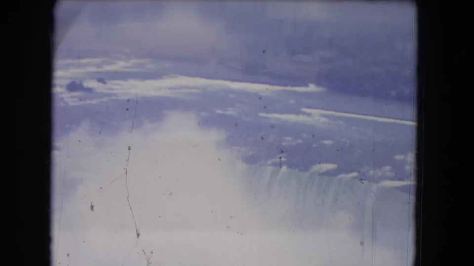 1964 8mm view of Niagara Falls with cascading water, broad mist cloud, blue-purple fading, and visible film gate shadows.