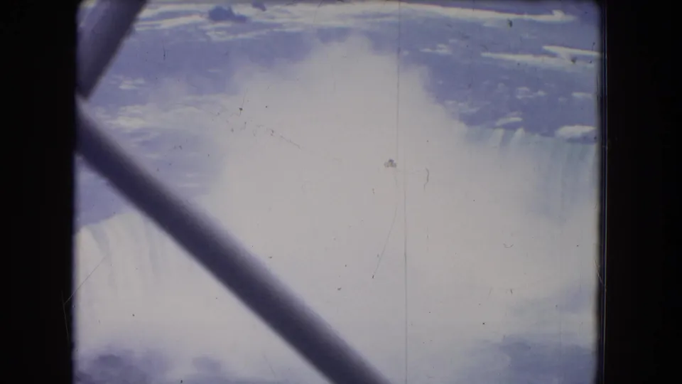 Elevated 1964 home-movie view of Niagara Falls, with heavy mist and a diagonal foreground support across the scene.