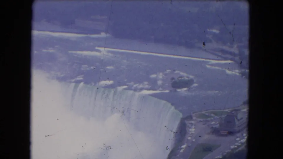 Aerial 8mm frame of Niagara Falls with the curved drop, mist, river, and nearby shoreline visible through dark supports.