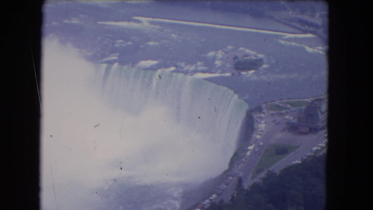 1964 Niagara Falls aerial frame with Horseshoe Falls, plume-like mist, river surface, and nearby roadway visible.