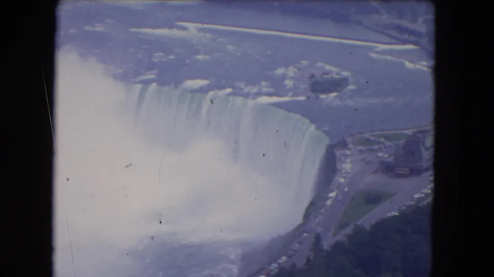 1964 Niagara Falls aerial frame with Horseshoe Falls, plume-like mist, river surface, and nearby roadway visible.
