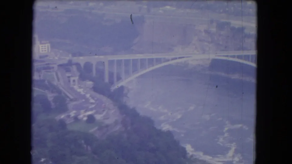 1964 8mm view near Niagara Falls showing mist, river, shoreline buildings and a wide arched bridge.