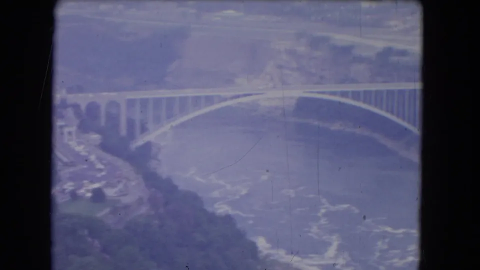 1964 8mm aerial view near Niagara Falls, New York, showing an arched bridge over a broad river with distant banks.