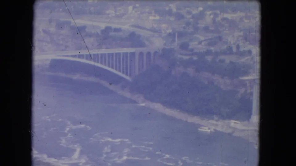 1964 8mm aerial footage near Niagara Falls, New York, showing the bridge at an angle with riverbank and water below.