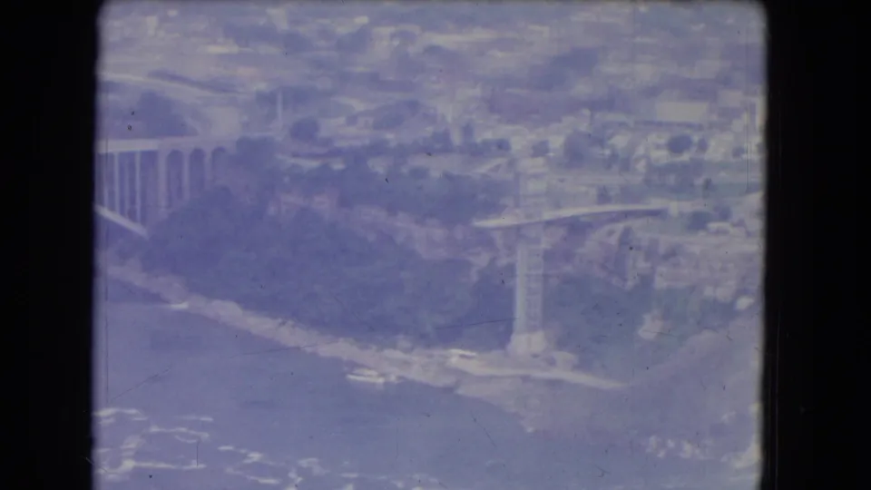 1964 8mm aerial view near Niagara Falls, New York, with bridge supports, river, shoreline, roads, and distant buildings.