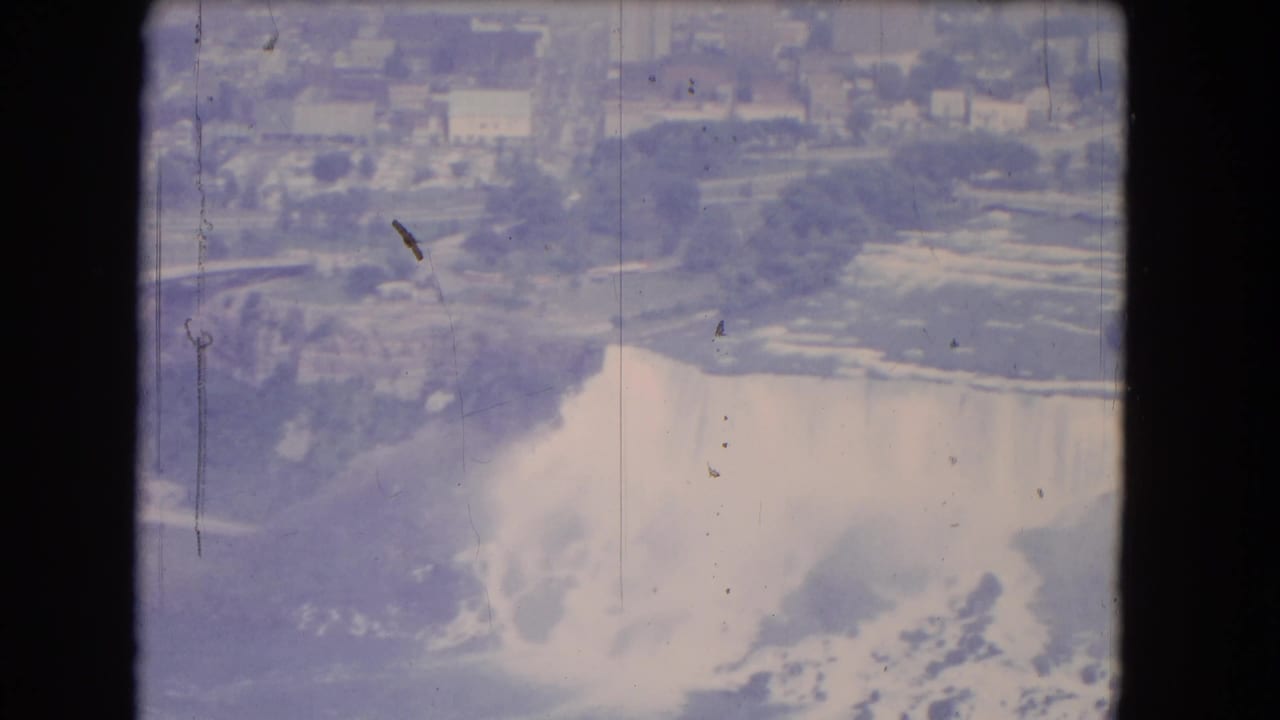 Aerial 8mm frame of American Falls and nearby Niagara Falls shoreline, with mist, roads, buildings, and greenery visible beyond the cataract.