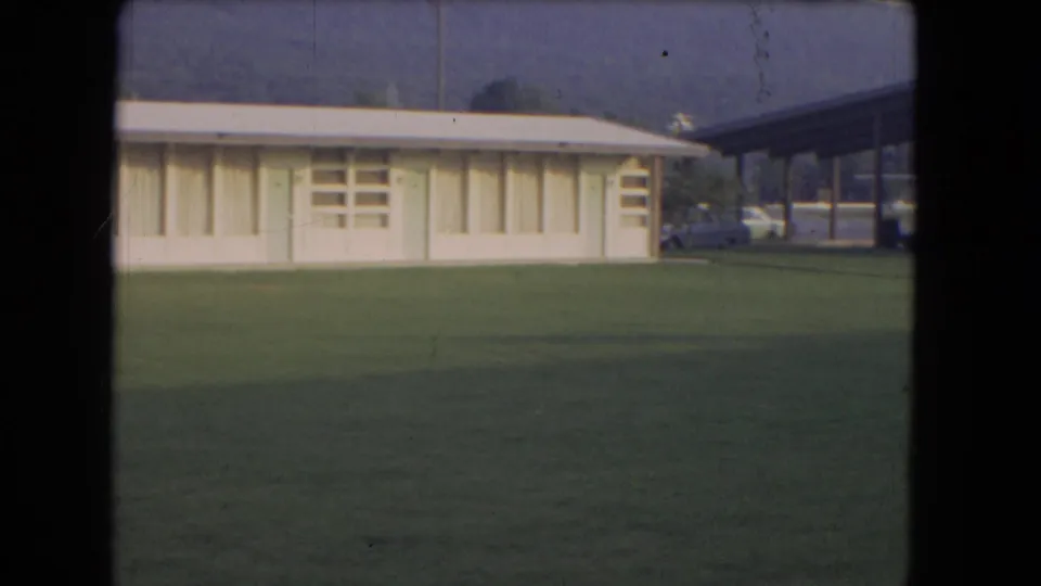 Ground-adjacent 1964 8mm view near Niagara Falls showing a low visitor-area building, green lawn, parked cars, hillside, and distant mist.