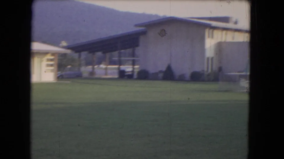 1964 8mm frame near Niagara Falls with a low modernist building, broad lawn, parked cars, hillside backdrop, and pale waterfall mist in the distance.
