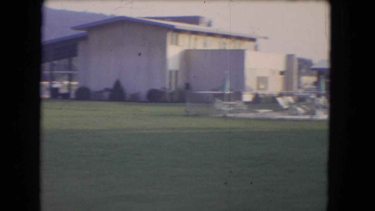 Poolside lawn with a low modernist building, lounge chairs, and closed umbrellas in soft 8mm color.