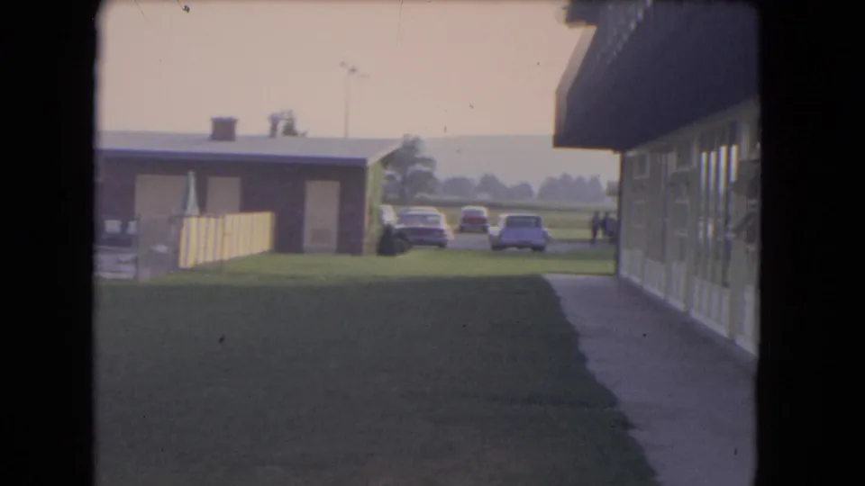 1964 Niagara Falls home-movie frame showing lawn, pool building, walkway, fence, parked cars, and distant people.