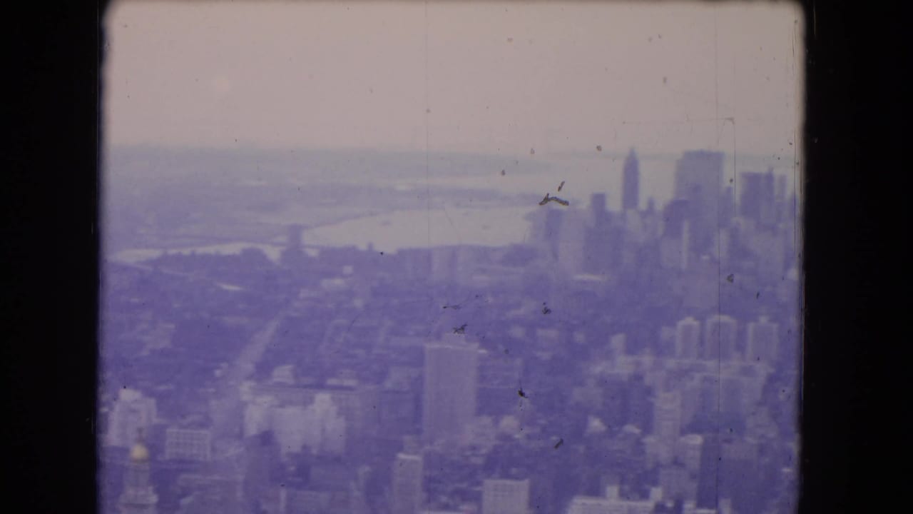 1964 8mm frame showing a hazy aerial cityscape with distant skyscrapers, waterfront, and dense urban blocks.