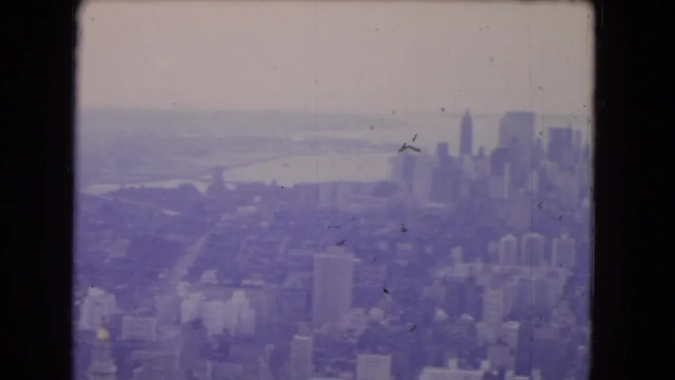 1964 8mm frame showing a hazy aerial cityscape with distant skyscrapers, waterfront, and dense urban blocks.