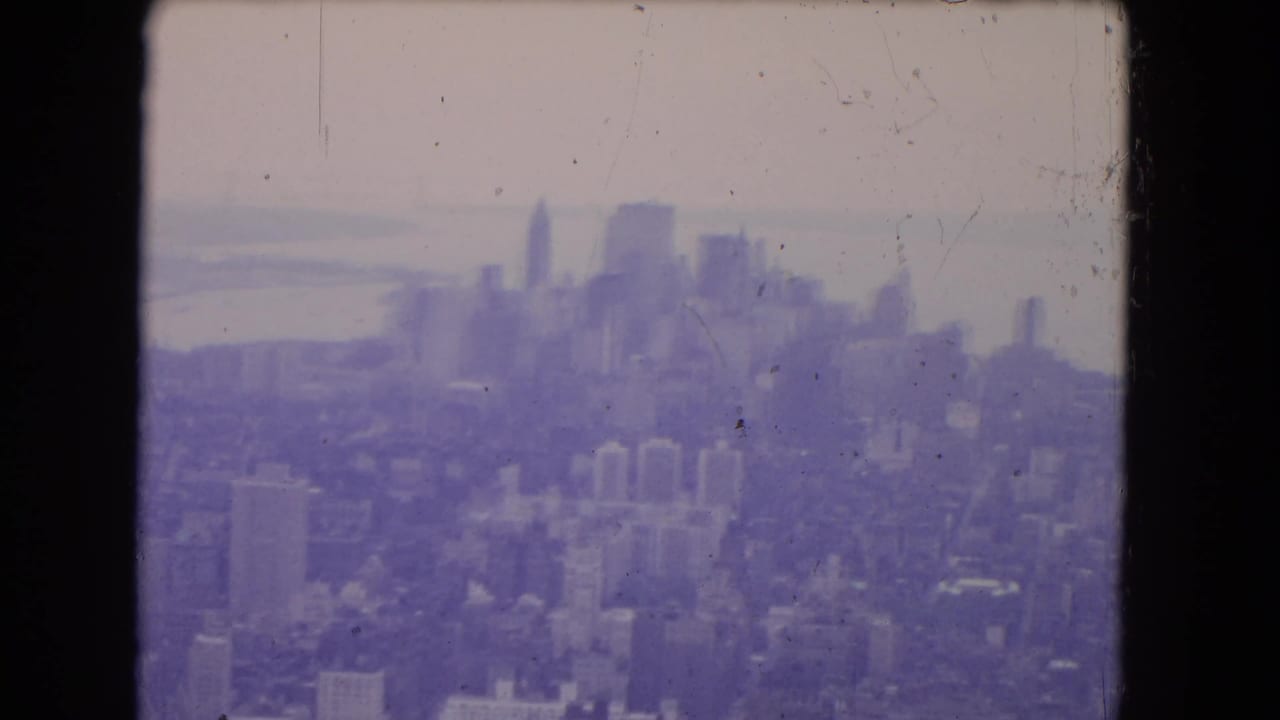 1964 8mm aerial frame with a soft urban skyline, waterfront haze, low-rise buildings, and visible film wear.