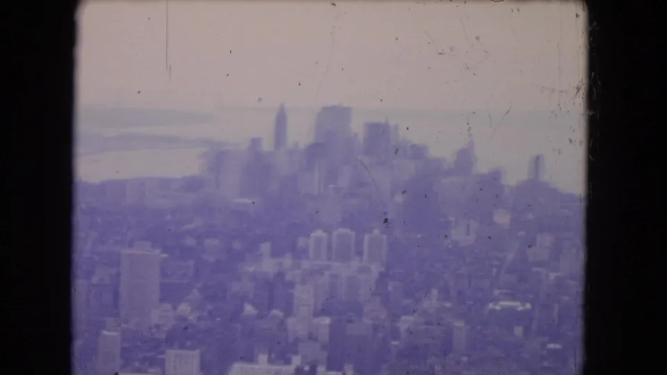 1964 8mm aerial frame with a soft urban skyline, waterfront haze, low-rise buildings, and visible film wear.