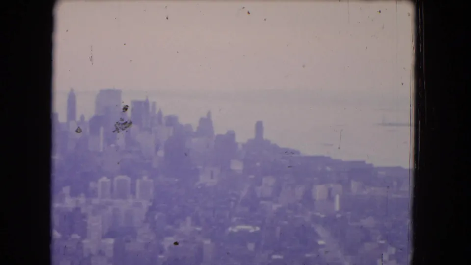 1964 8mm frame showing a blurred cityscape from above, with tall buildings, dense blocks, and water in the distance.