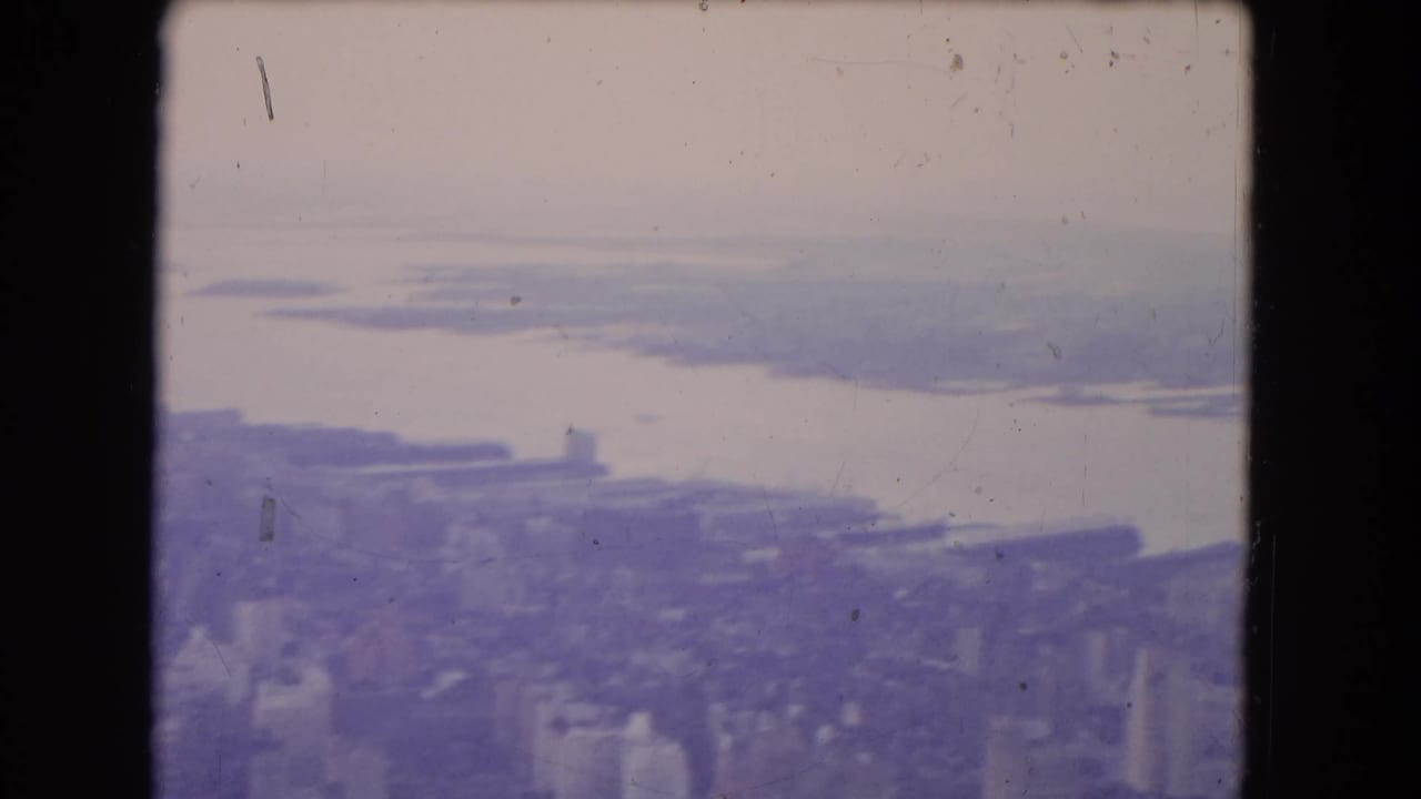 High aerial view over dense city blocks beside a broad body of water.