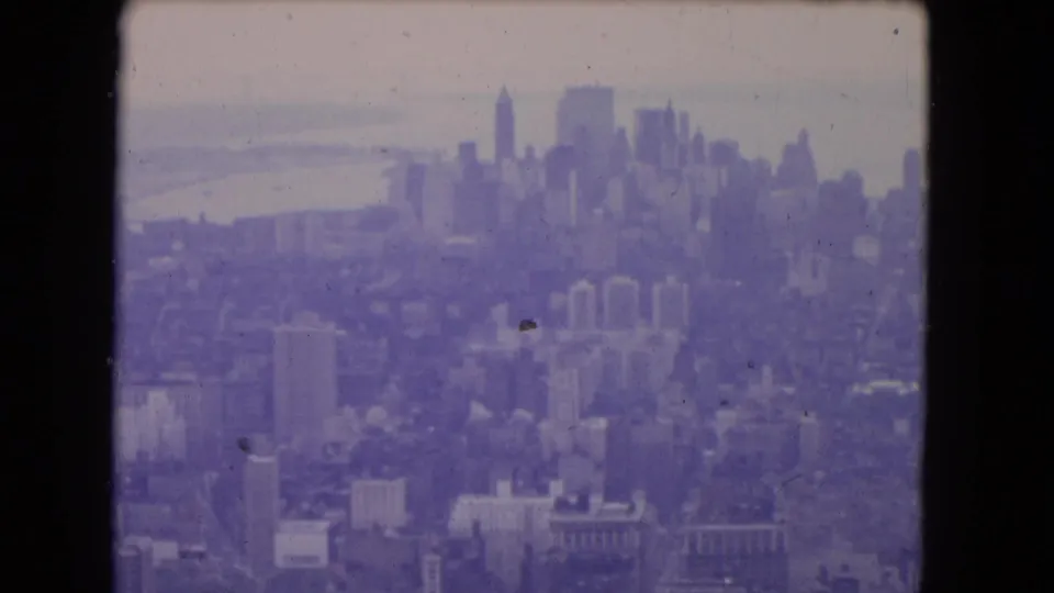 1964 8mm frame of the New York City skyline, with tall buildings beyond lower blocks and water visible at left.