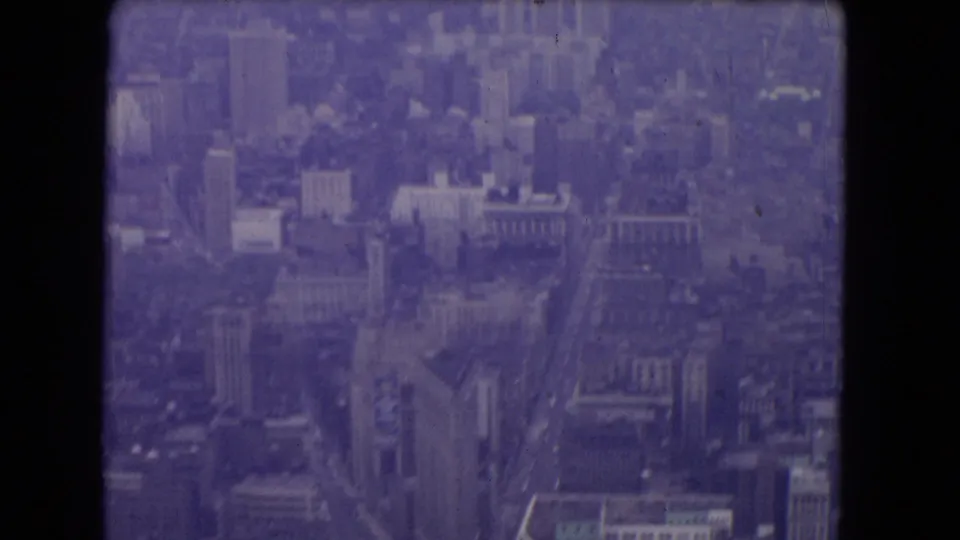 1964 8mm frame showing a broad aerial look over New York City buildings, streets, and waterfront distance with visible film scratches.