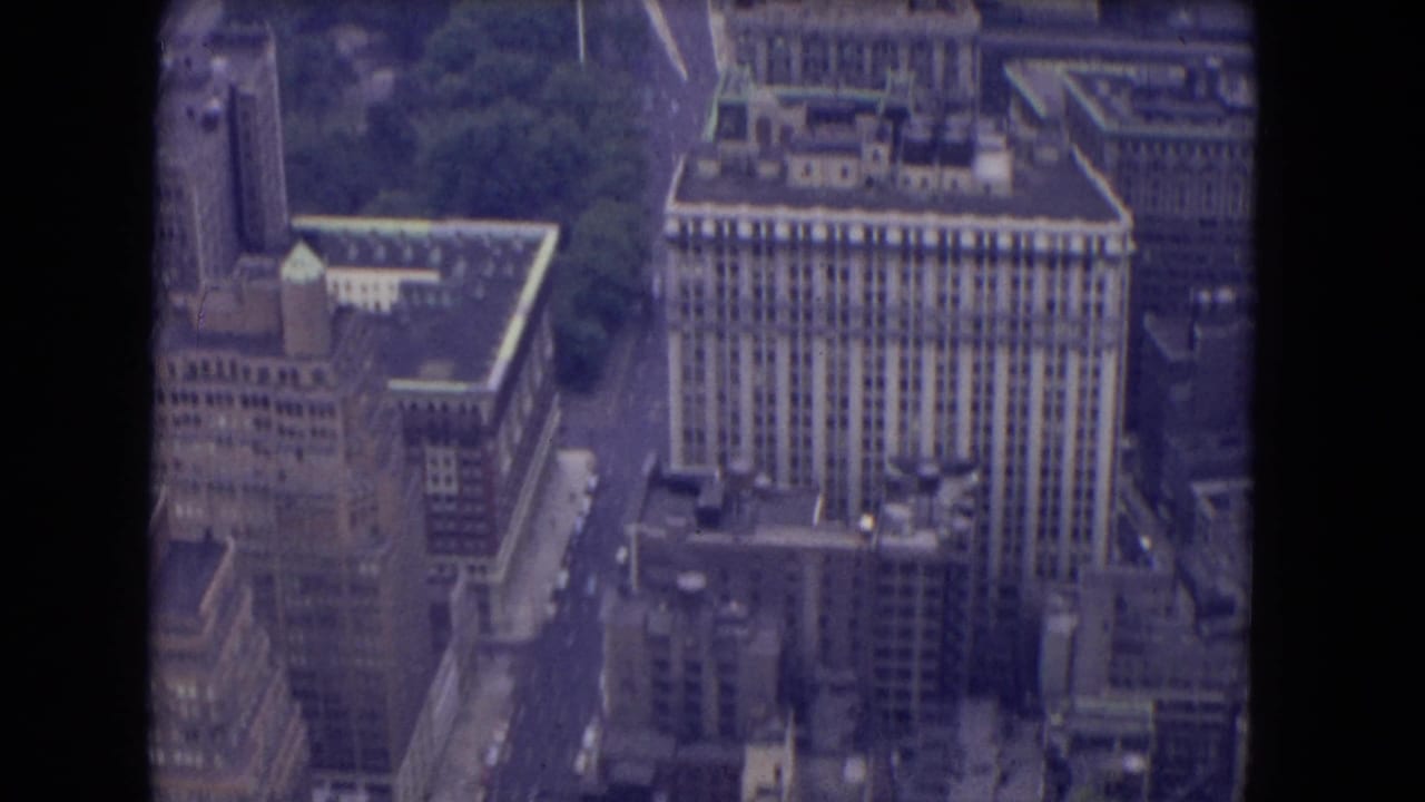Elevated 8mm city view with a large rectangular high-rise, older masonry buildings, narrow streets, rooftops, and soft purple film color.