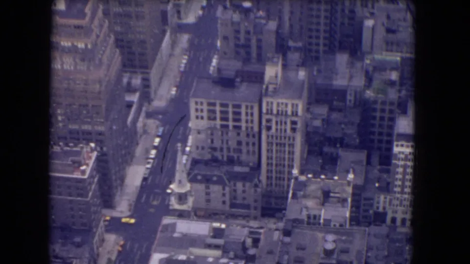 1960s 8mm aerial view over downtown blocks, showing tall buildings, a church-like spire, roof surfaces, streets, and small cars.