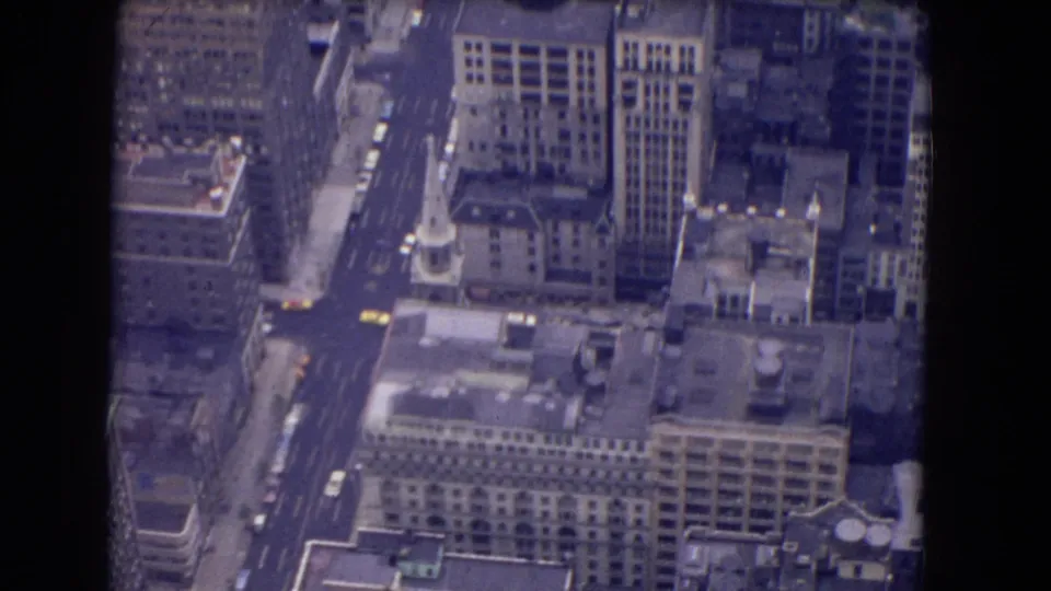 Elevated 8mm view of a dense urban district with high-rises, a church-like spire, rooflines, streets, and scattered cars below.