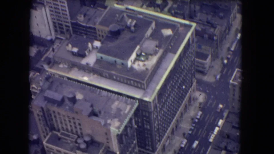 1964 8mm aerial frame showing a large high-rise, rooftop equipment, nearby streets, and small cars seen from above.
