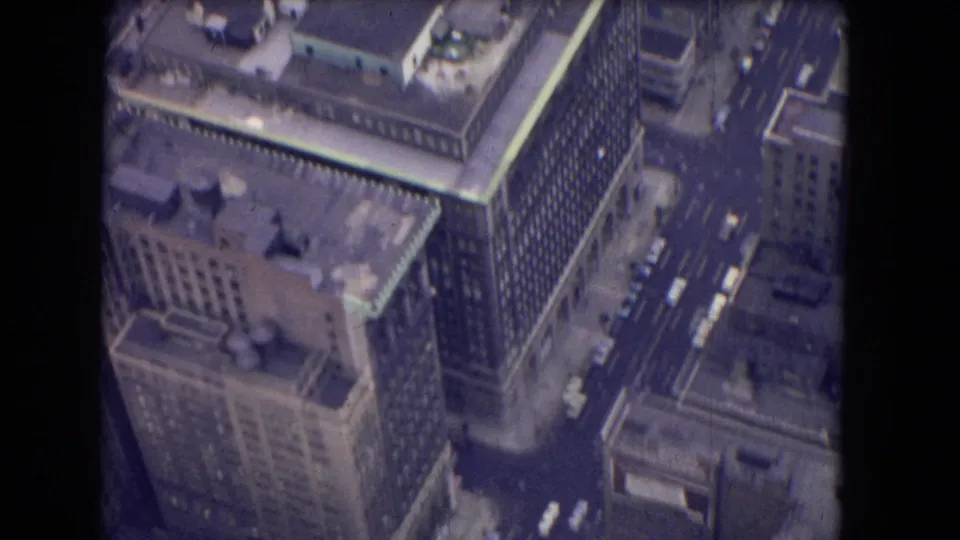 1964 8mm aerial view of tall urban buildings with flat roofs, street traffic, and a dark film-frame edge visible.