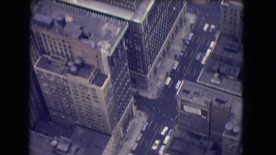 1964 8mm aerial frame with a large high-rise, adjacent buildings, gridded streets, and cars visible from above.