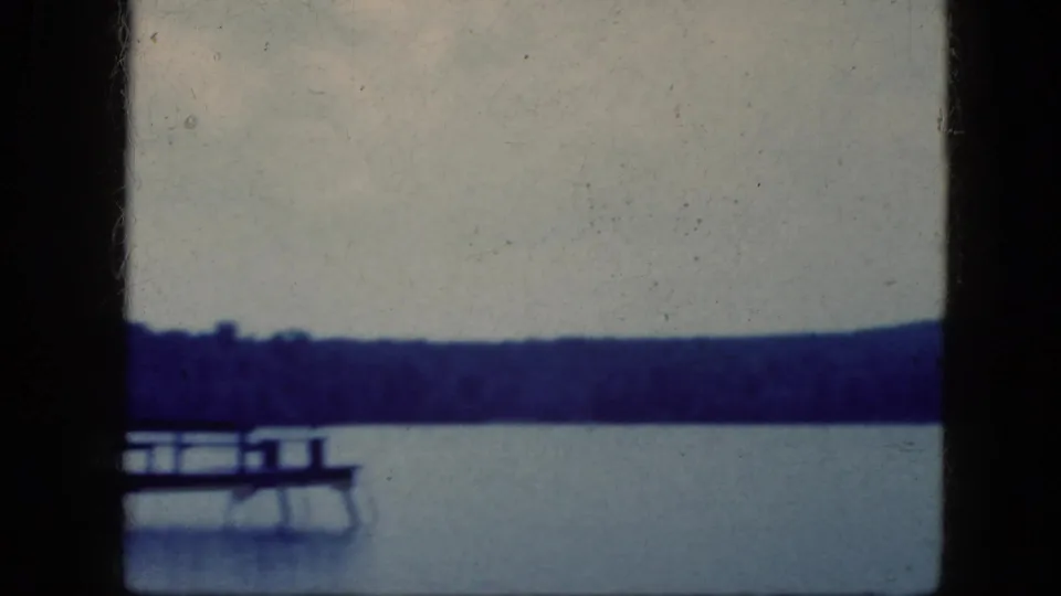 1964 lakeside home-movie frame showing a wooden dock and a small boat against a distant tree-lined shore.