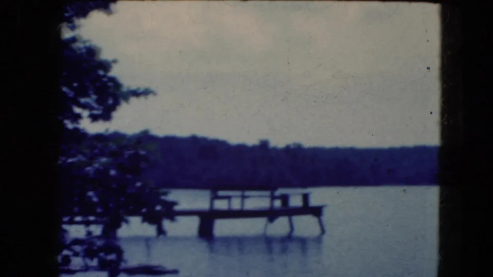 1964 8mm frame from a lakeside viewpoint, showing a wooden dock, nearby tree branches, calm water, and distant woods.