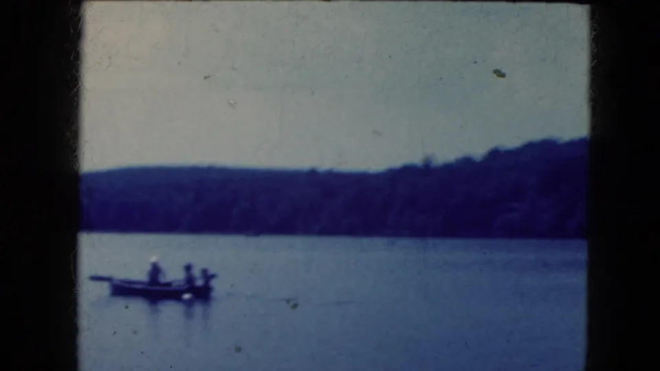 1964 home-movie frame showing a small boat with distant people on calm water near a wooded shoreline in New York metadata context.