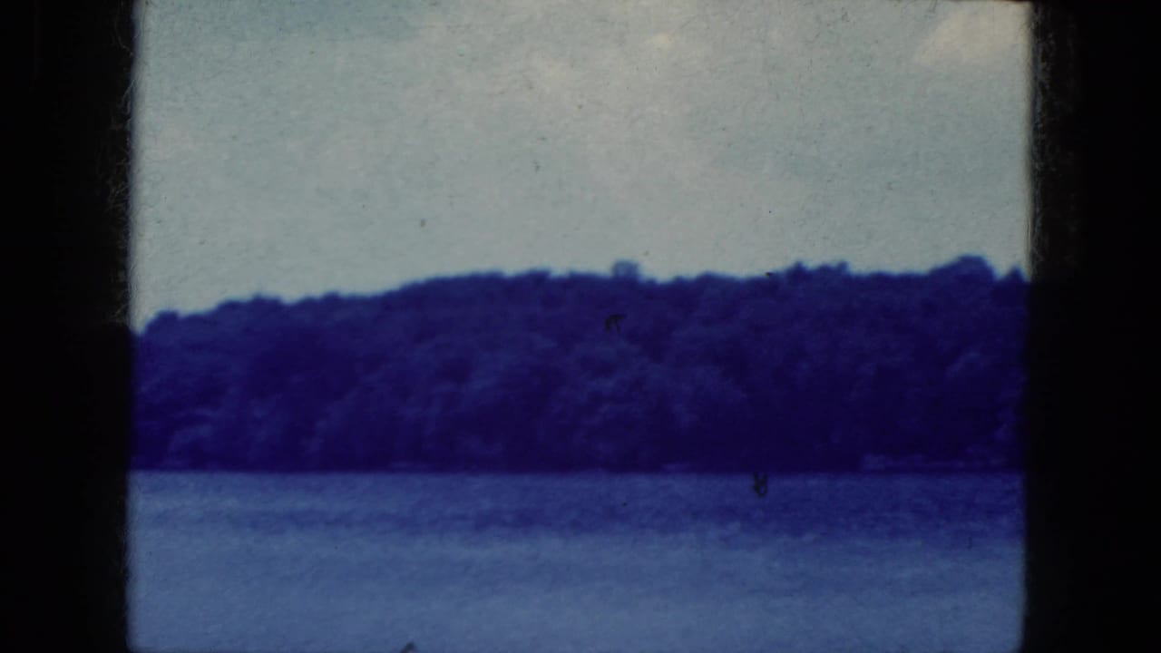 Static 1964 8mm view of a lake and wooded shoreline near Niagara Falls, New York, with blue color shift and visible frame edge.