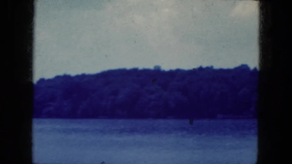 Static 1964 8mm view of a lake and wooded shoreline near Niagara Falls, New York, with blue color shift and visible frame edge.