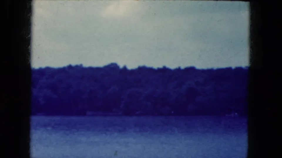 1964 home-movie frame showing water and a distant forested shoreline in the Niagara Falls, New York area, softened by grain and blue tint.