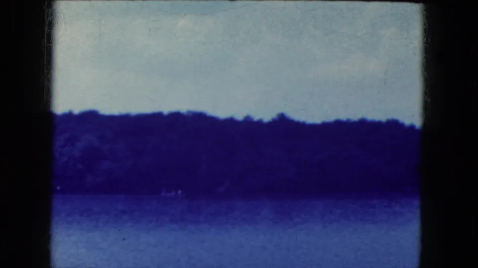 End frame of a 1964 8mm lakeside view near Niagara Falls, New York, with calm water, dark tree line, pale sky, and gate edge.