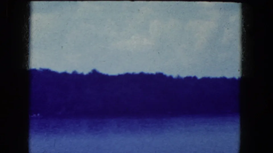 1964 8mm home-movie frame from Niagara Falls, New York, showing calm water, a distant wooded shore, and a pale sky with heavy blue tint.