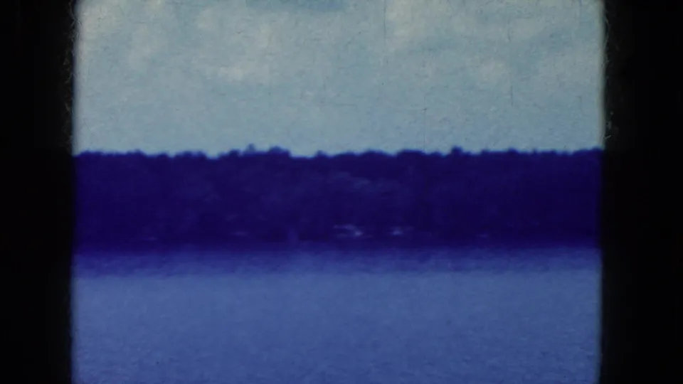 1964 8mm travel footage frame showing a blue-tinted lake or river surface, dark tree line, pale cloudy sky, and visible film texture.