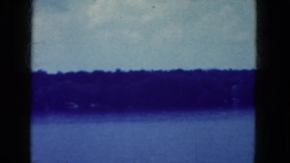 Blue-tinted 8mm frame from the Niagara Falls, New York clip, showing calm water, a low distant tree line, pale sky, scratches, and film borders.