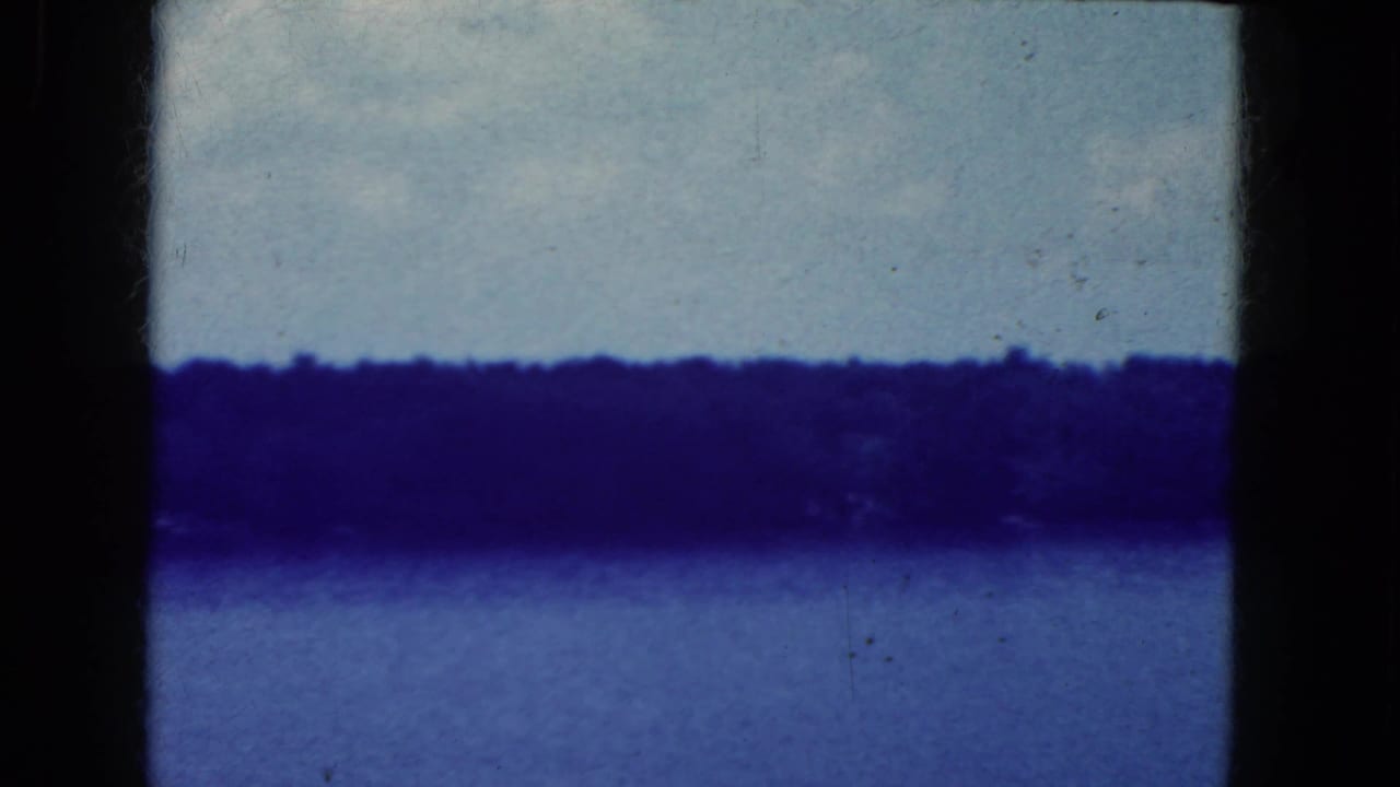 1964 8mm home-movie frame of a calm lake near Niagara Falls, New York metadata, with a dark shoreline under pale sky.
