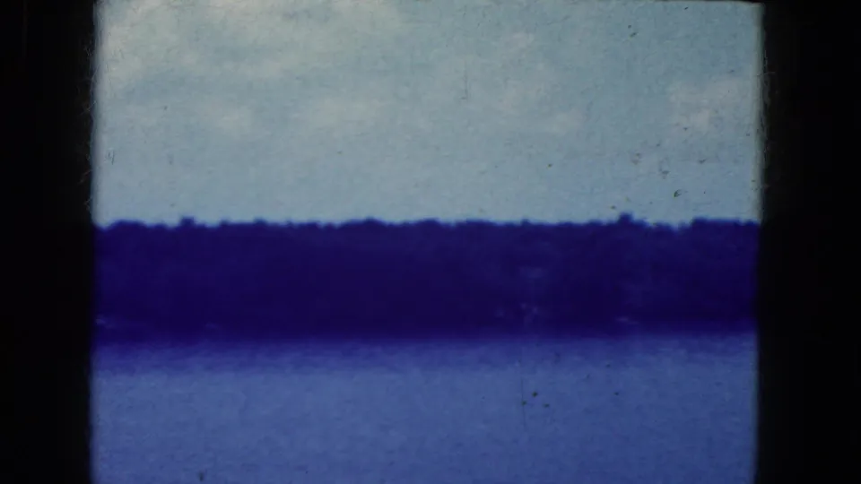 1964 8mm home-movie frame of a calm lake near Niagara Falls, New York metadata, with a dark shoreline under pale sky.