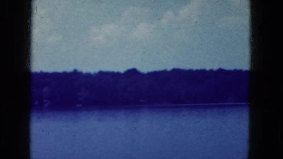 Blue-tinted 8mm frame showing lake, distant wooded shore, pale sky, and a dark vertical divider from the contact sheet.