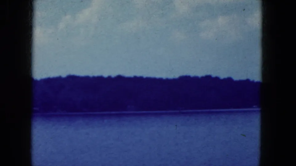 1964 home-movie still of a blue-toned lakeside view, with wooded shoreline, sky, film grain, and contact-sheet borders.