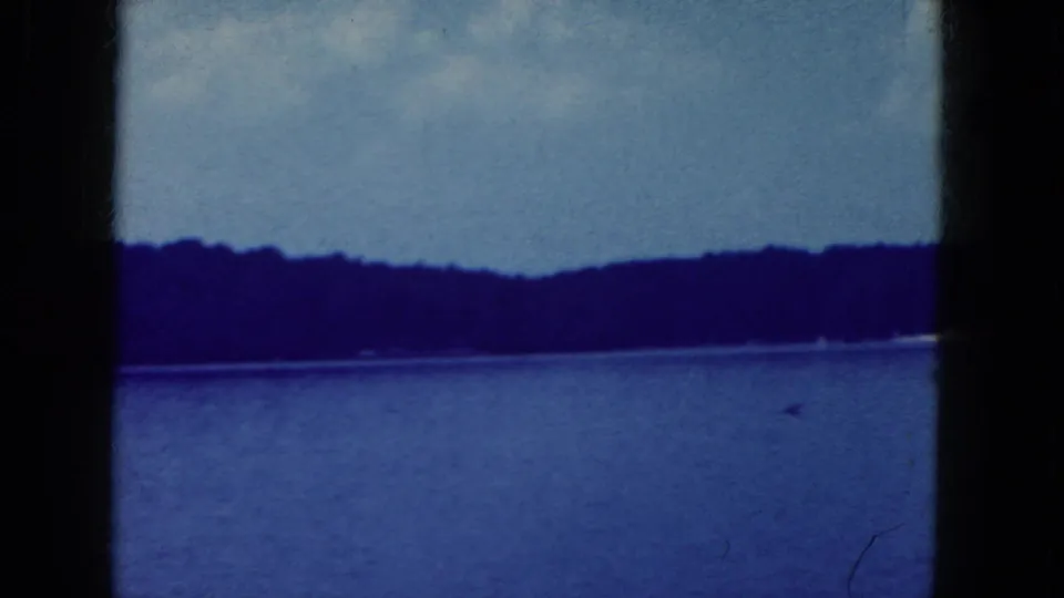 1964 8mm frame from the Niagara Falls, New York area showing calm lake water, distant wooded shoreline, and dark gate borders.