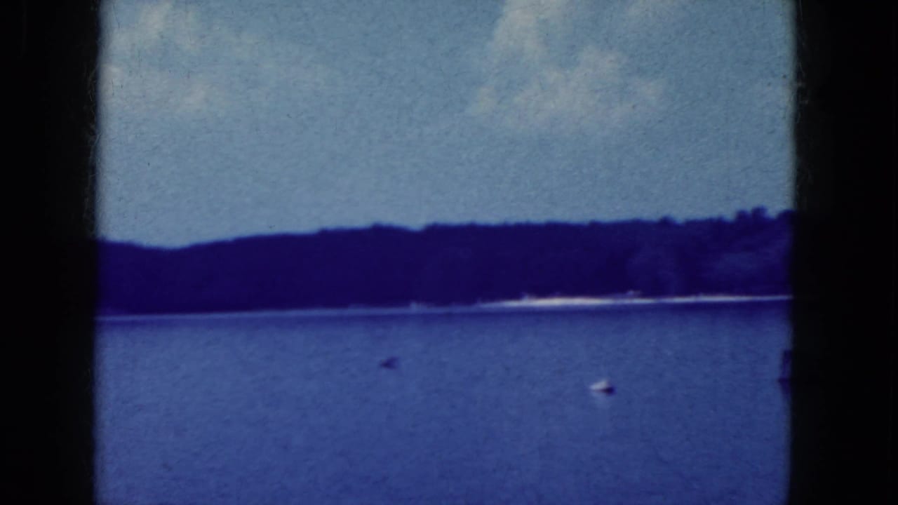 1964 8mm lakeside view near Niagara Falls, New York, with a wooded horizon, blue water, and a small white buoy or boat.