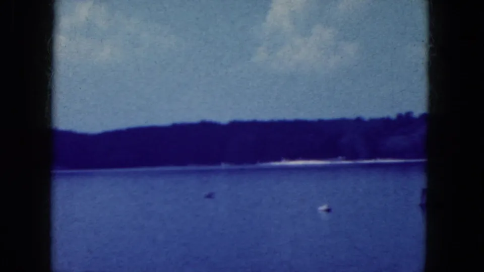 1964 8mm lakeside view near Niagara Falls, New York, with a wooded horizon, blue water, and a small white buoy or boat.