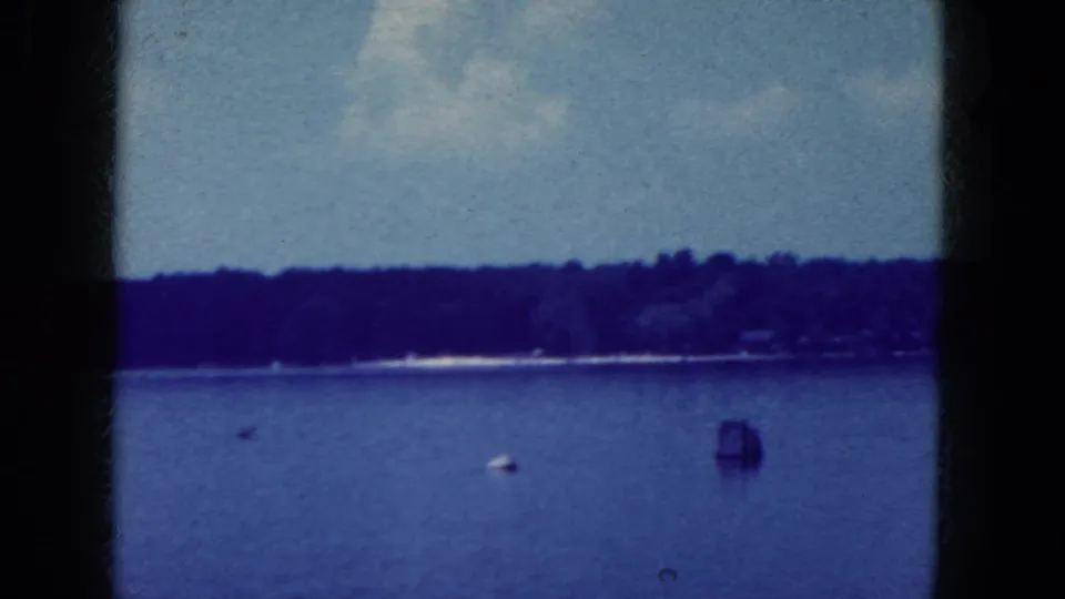 1964 8mm frame of a recreational lake near Niagara Falls, New York, with water, shoreline, and small objects visible offshore.