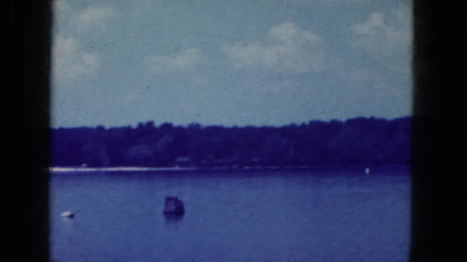 1964 8mm lakeside view near Niagara Falls, New York, showing a tree-lined shore, blue water, and small waterborne objects.